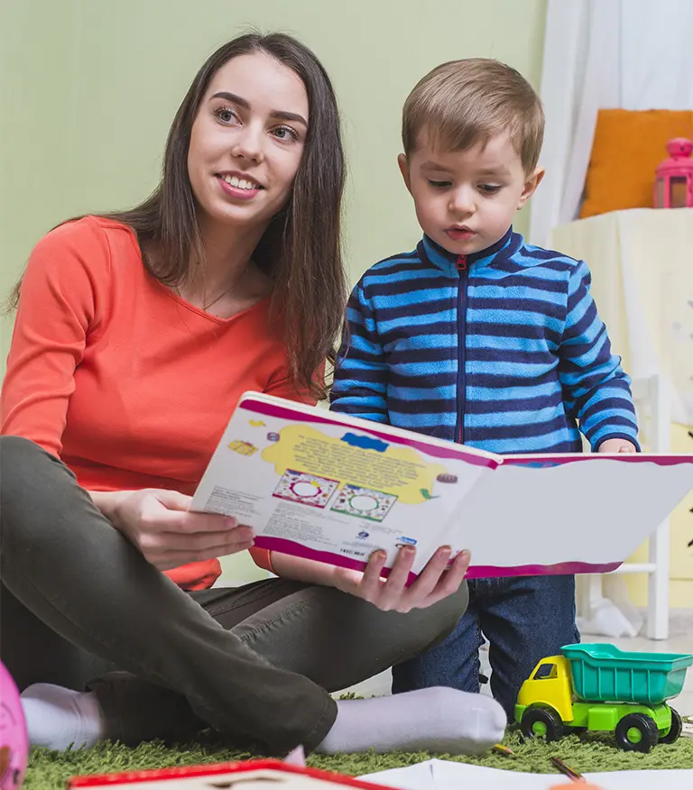 Smiling early childhood education apprentice reading a book to a young boy in a childcare setting in France